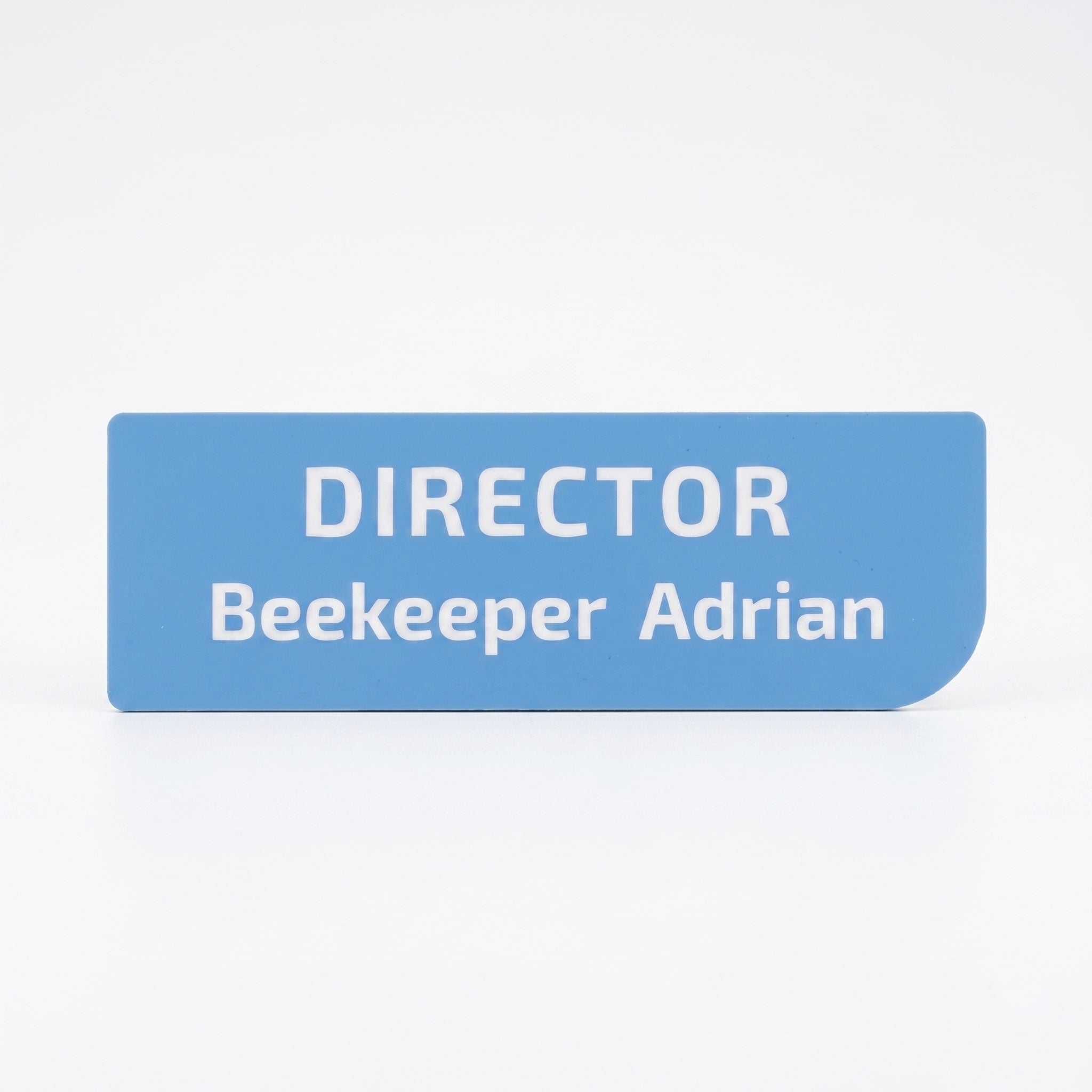 Plăcuță de identificare simplă pentru birou din acril albastru deschis mat, cu colțul din dreapta jos rotunjit asimetric, gravată cu textul alb „DIRECTOR Beekeeper Adrian”, pe fundal alb.