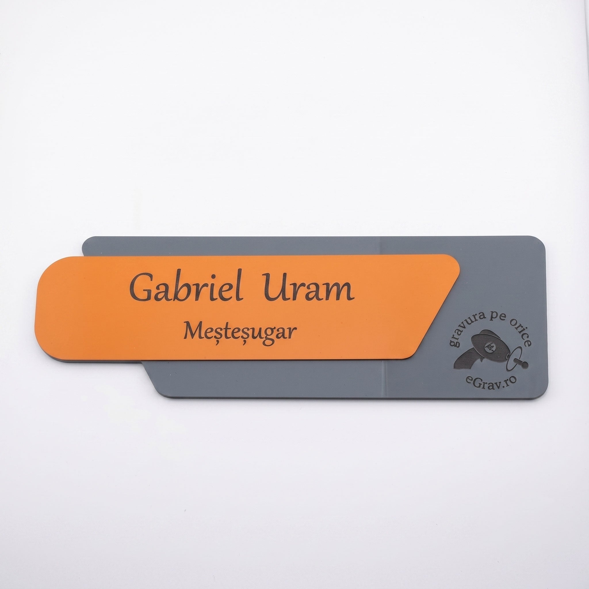 Plăcuță de identificare asimetrică pentru birou din două elemente, cu fața portocaliu mat gravată cu numele „Gabriel Uram Meșteșugar” și o bază gri închis mat cu logo-ul eGrav.ro, pe fundal alb.