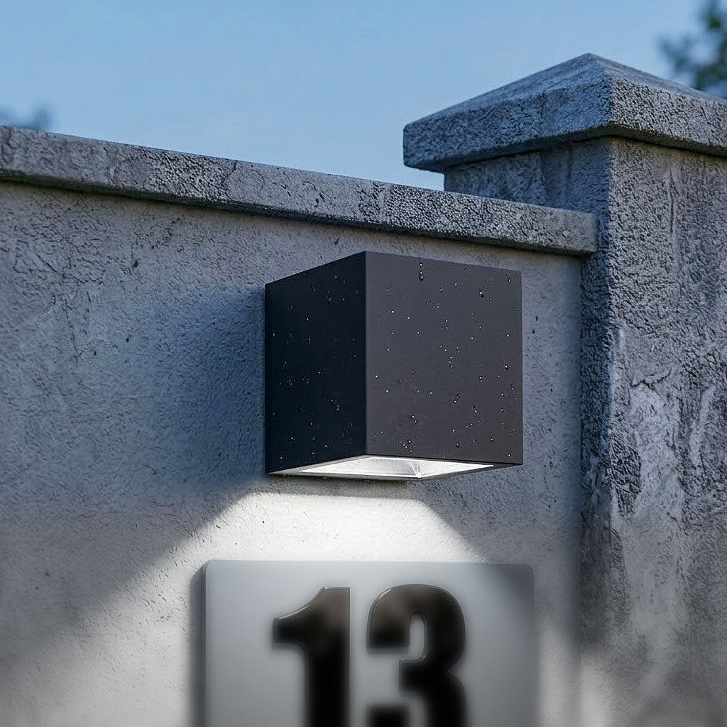 Aplica solara LED cubica neagra montata pe un gard modern din sipci de lemn orizontale si stalpi de beton. Lampa este orientata in jos si ilumineaza o placuta cu numarul de casa '15', intr-o curte interioara bine amenajata, la amurg.