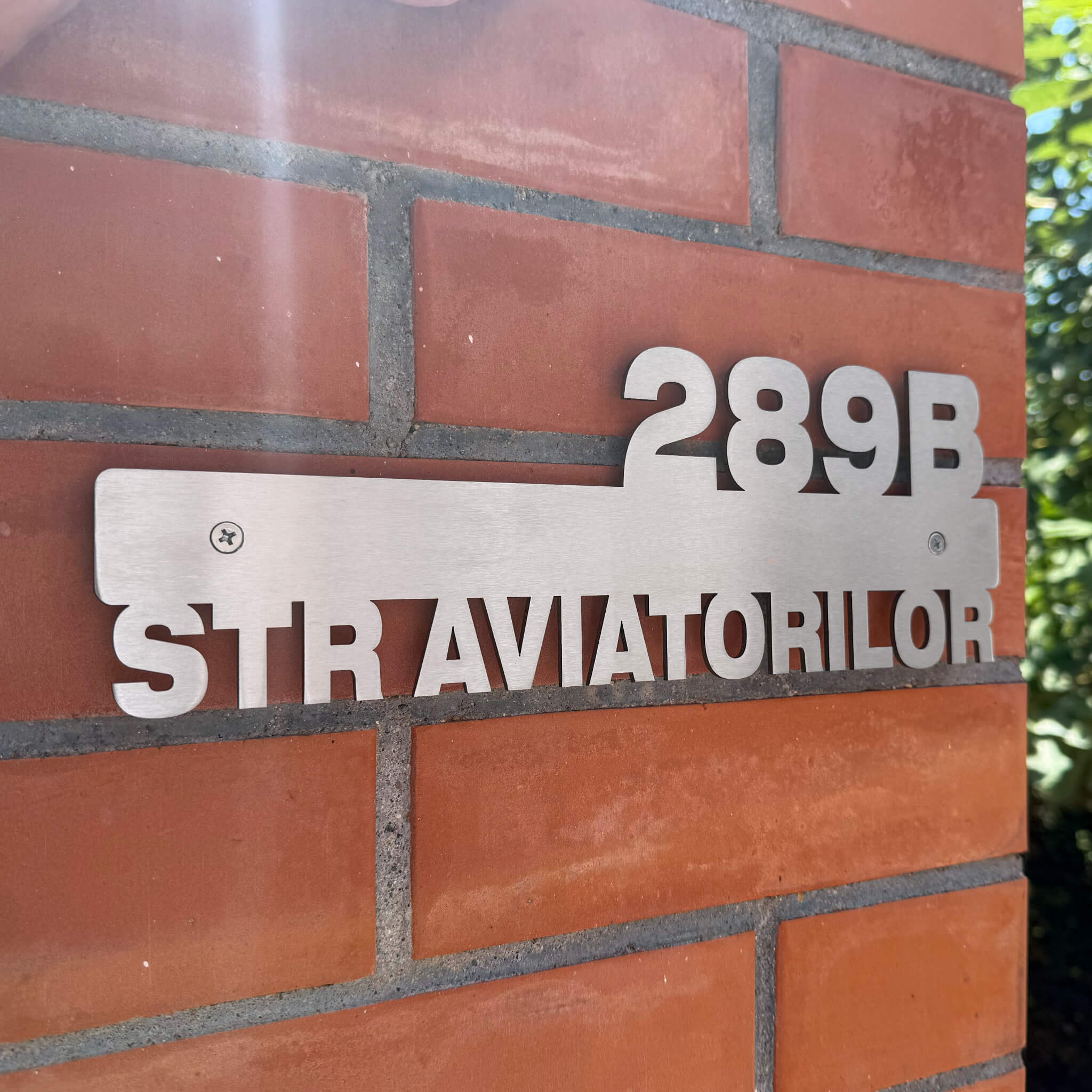 Plăcuță de adresă modernă din inox, personalizată prin decupare laser, fixată cu șuruburi pe un stâlp de cărămidă.