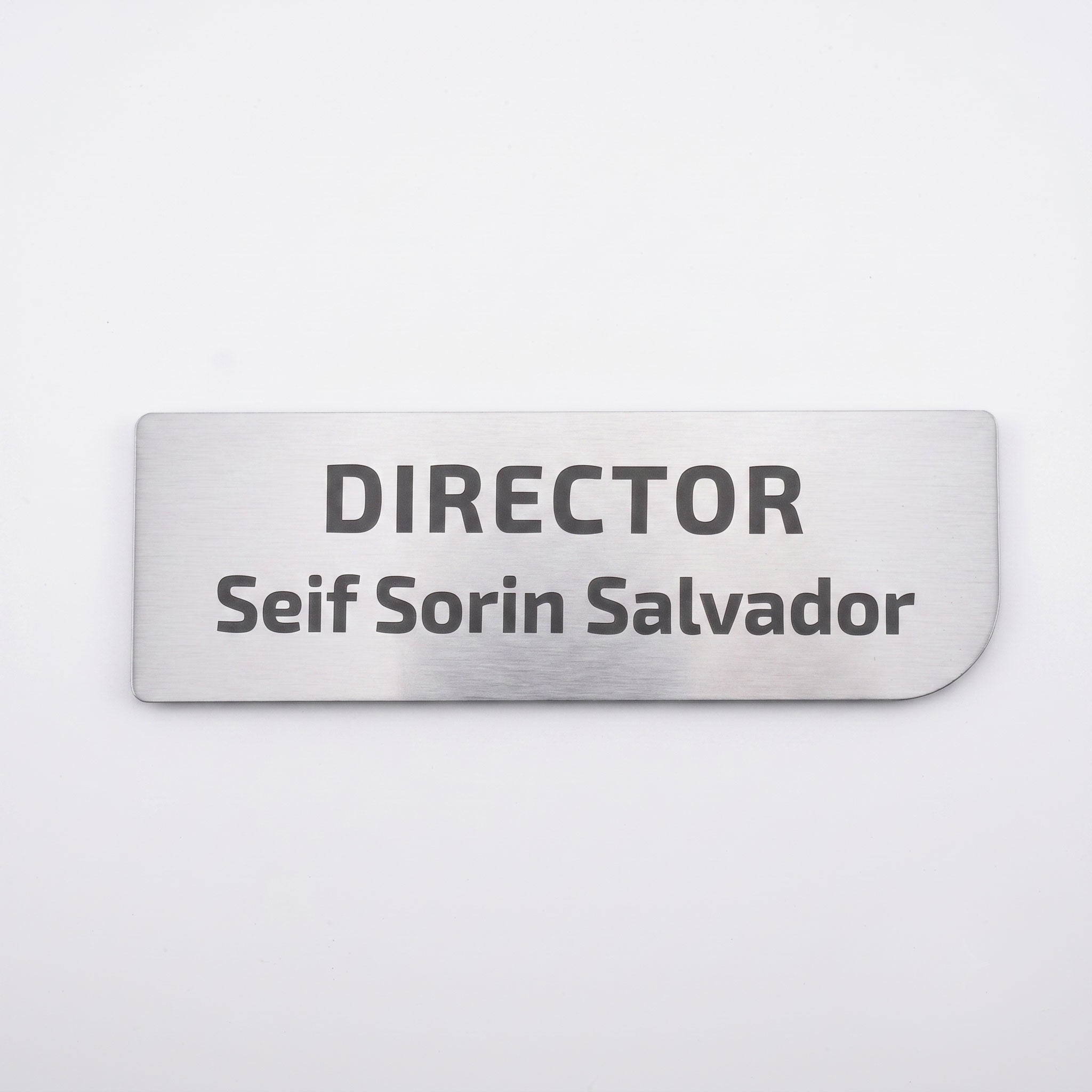 Plăcuță de identificare simplă pentru birou din metal argintiu periat, cu colțul din dreapta jos rotunjit asimetric, gravată cu textul „DIRECTOR Seif Sorin Salvador”, pe fundal alb.