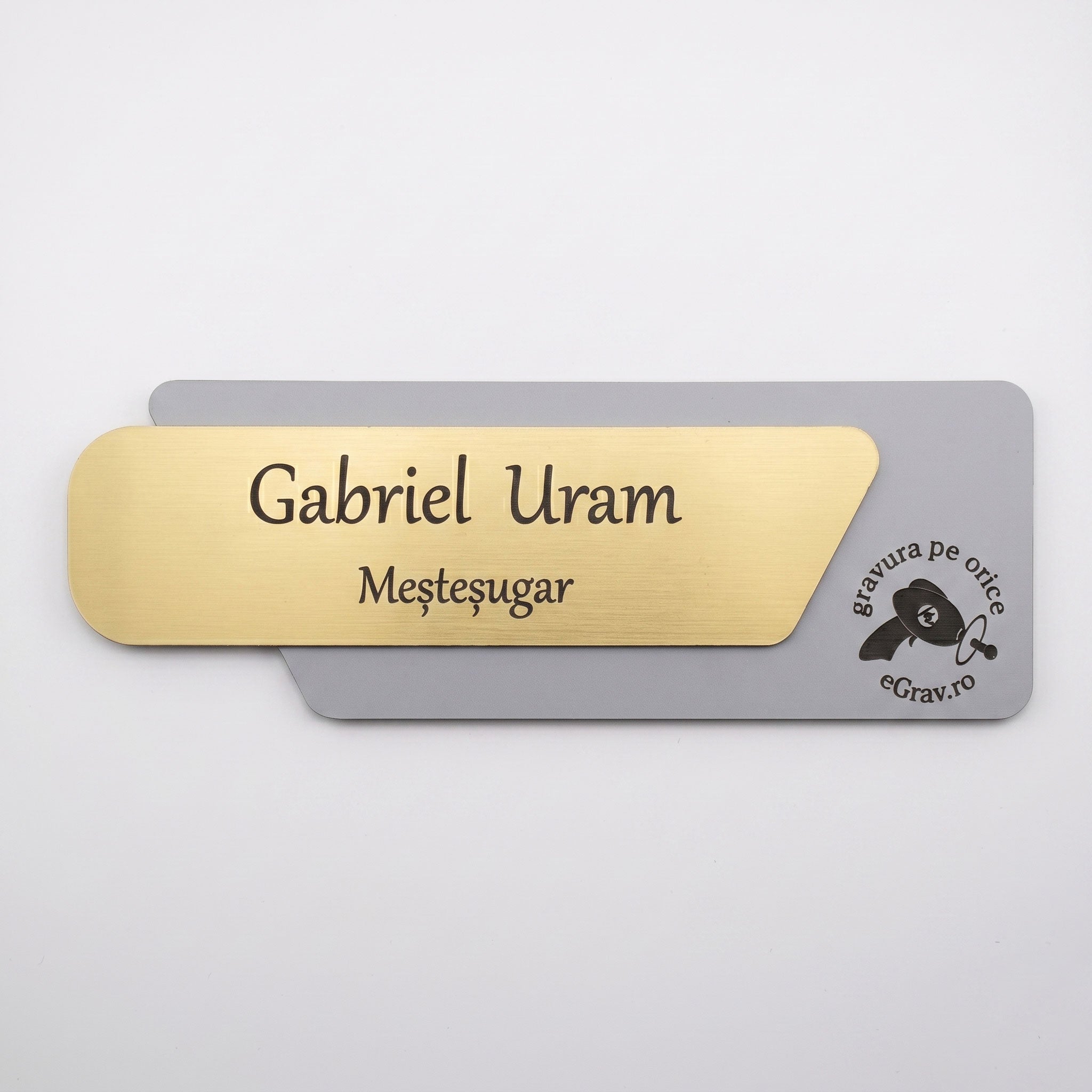 Plăcuță premium de identificare pentru birou din două elemente, cu fața asimetrică metalică auriu periat gravată cu textul „Gabriel Uram Meșteșugar” și o bază gri mat cu logo-ul eGrav.ro, pe fundal alb.