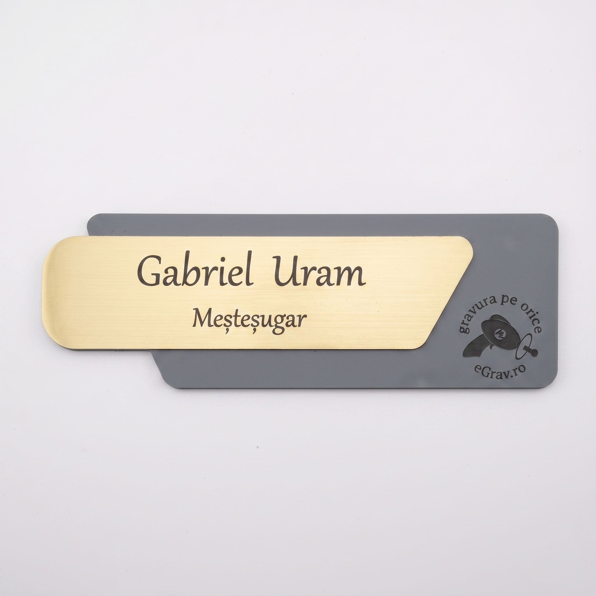 Plăcuță de identificare asimetrică pentru ușă de birou formată din două elemente, cu fața din metal auriu periat gravată „Gabriel Uram Meșteșugar” și baza gri închis mat cu logo-ul eGrav.ro, pe fundal alb.