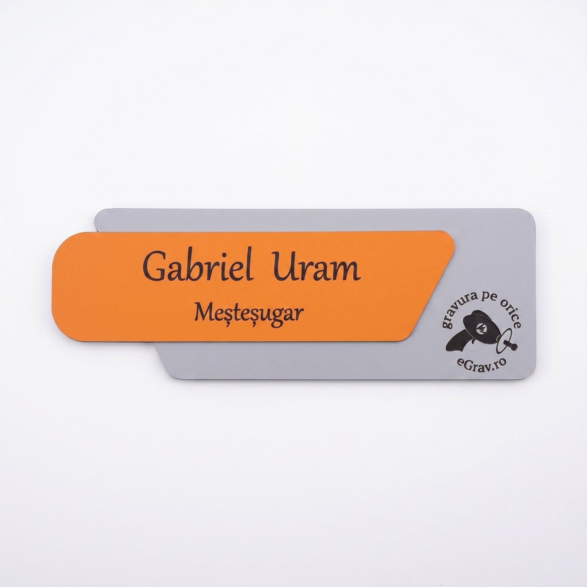 Plăcuță de identificare asimetrică pentru birou formată din două elemente, cu fața portocaliu mat gravată cu textul „Gabriel Uram Meșteșugar” și baza gri mat cu logo-ul eGrav.ro, pe fundal alb.