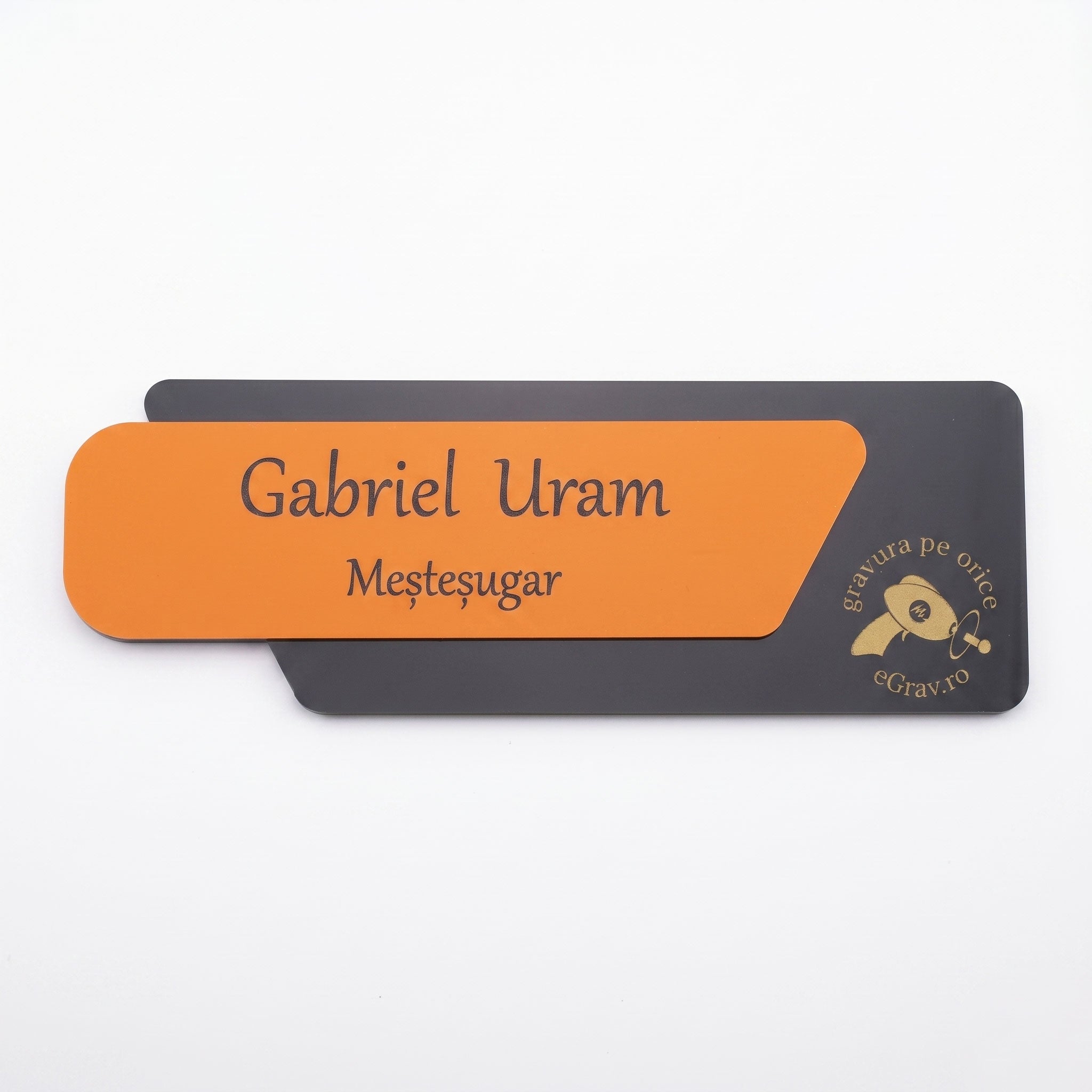 Plăcuță de identificare asimetrică pentru birou din două elemente, cu fața portocaliu mat gravată cu textul „Gabriel Uram Meșteșugar” și o bază gri închis mat cu logo-ul eGrav.ro, pe fundal alb.