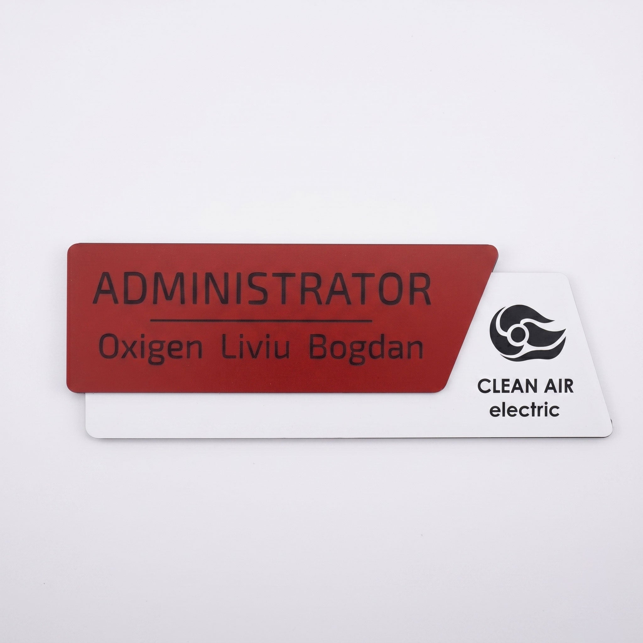 Plăcuță de identificare asimetrică pentru birou formată din două elemente, cu fața roșu închis mat gravată cu numele „ADMINISTRATOR Oxigen Liviu Bogdan” și o bază albă mat cu logo-ul Clean Air electric, pe fundal alb.