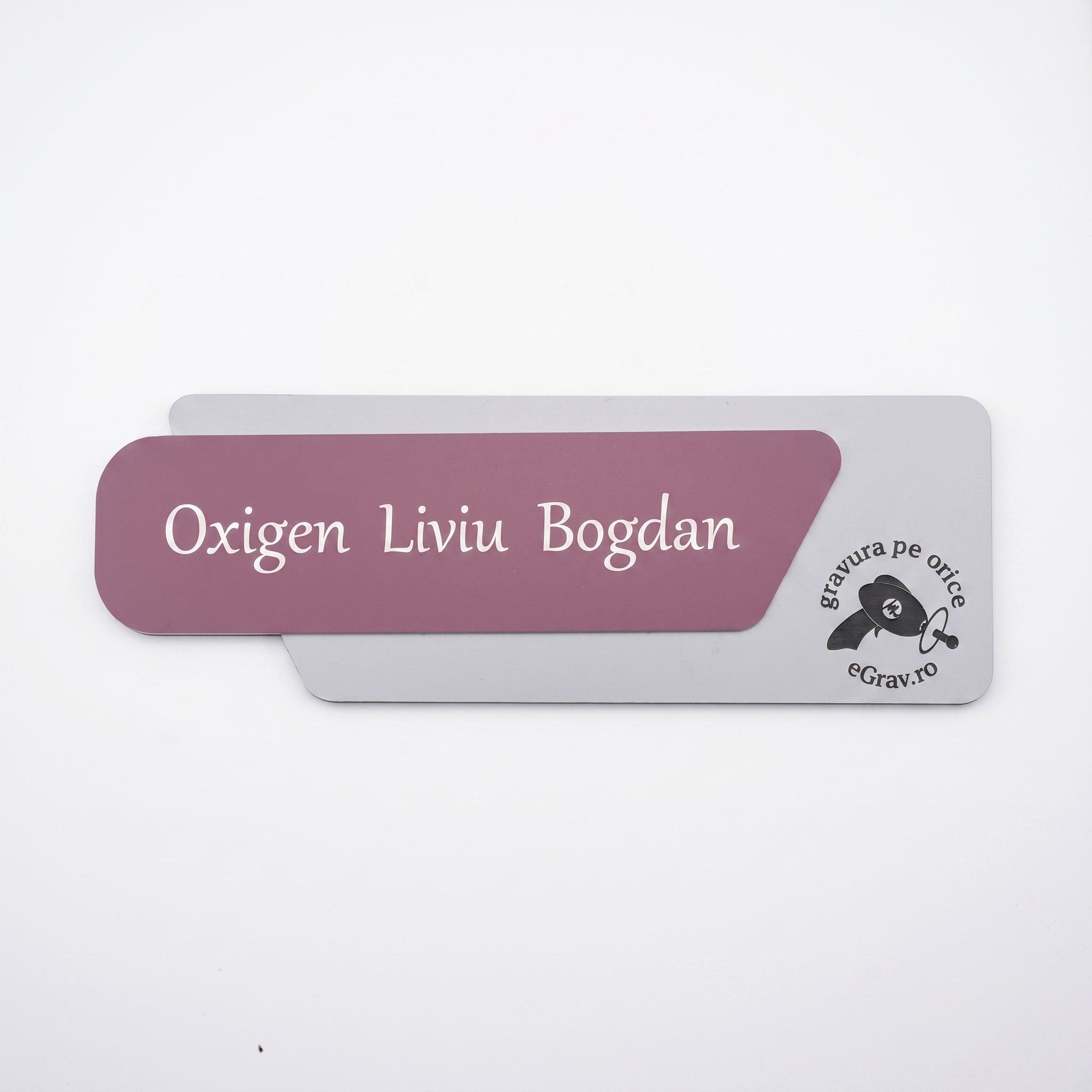 Plăcuță de identificare asimetrică pentru birou formată din două elemente, cu fața roz prăfuit mat gravată cu textul „Oxigen Liviu Bogdan” și baza gri mat cu logo-ul eGrav.ro, pe fundal alb.