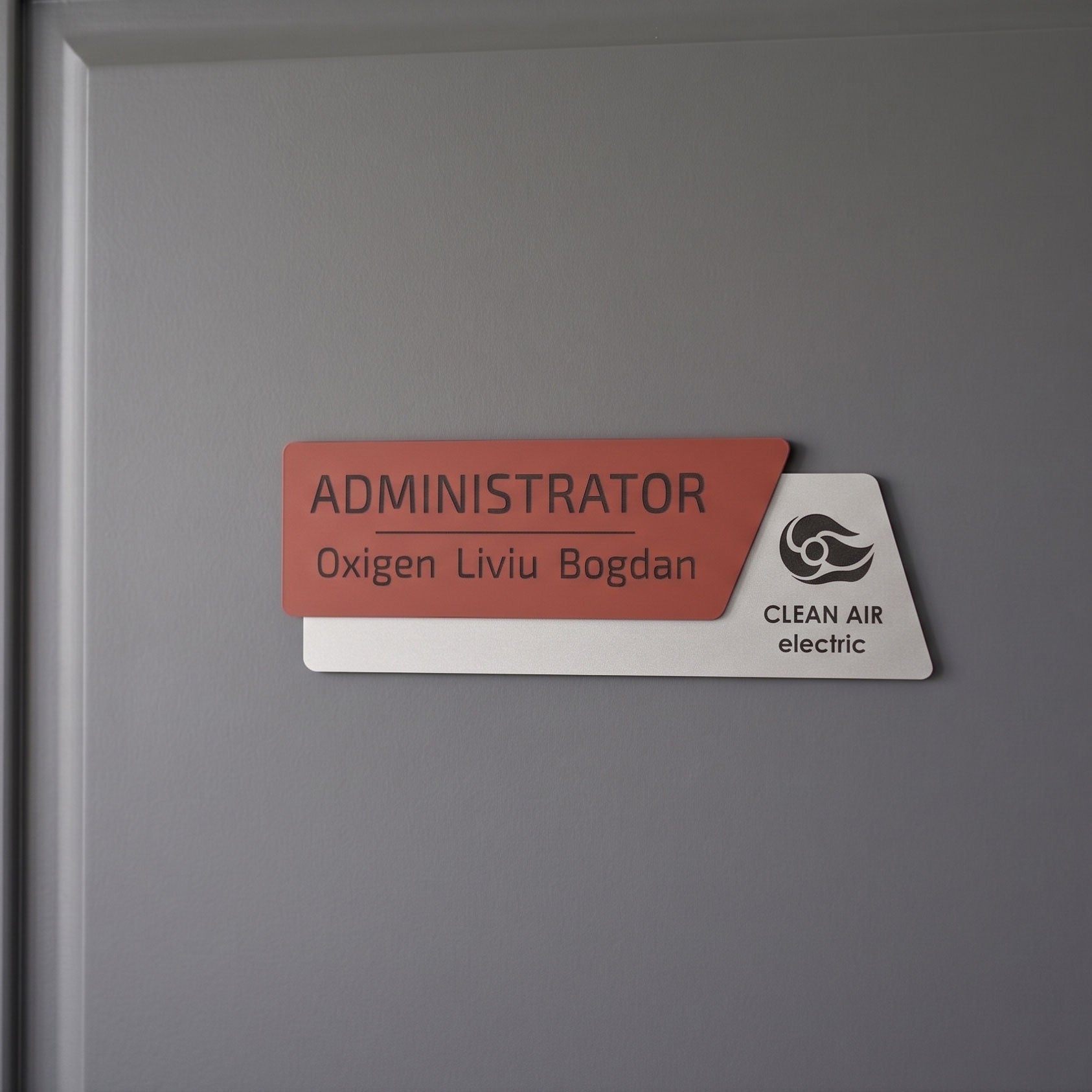 Plăcuță de identificare asimetrică pentru birou formată din două elemente, cu fața roșu închis mat gravată cu numele „ADMINISTRATOR Oxigen Liviu Bogdan” și o bază albă mat cu logo-ul Clean Air electric, pe fundal alb.