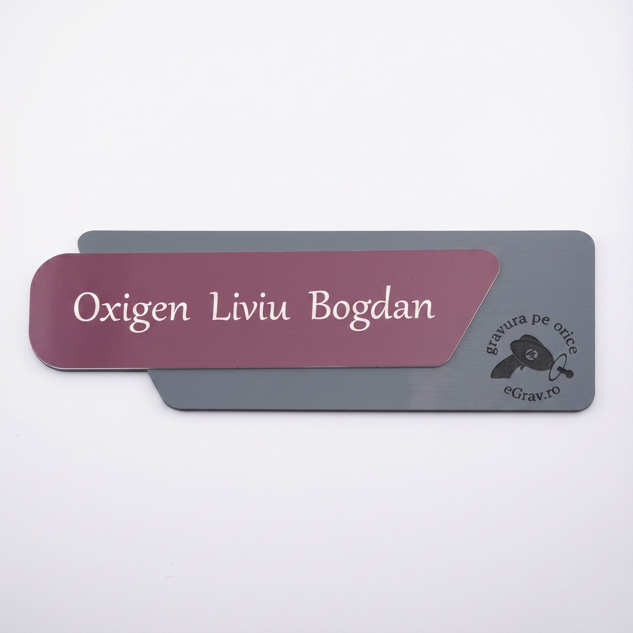 Plăcuță de identificare asimetrică pentru birou formată din două elemente, cu fața roz prăfuit mat gravată cu textul „Oxigen Liviu Bogdan” și o bază gri închis mat cu logo-ul eGrav.ro, pe fundal alb.