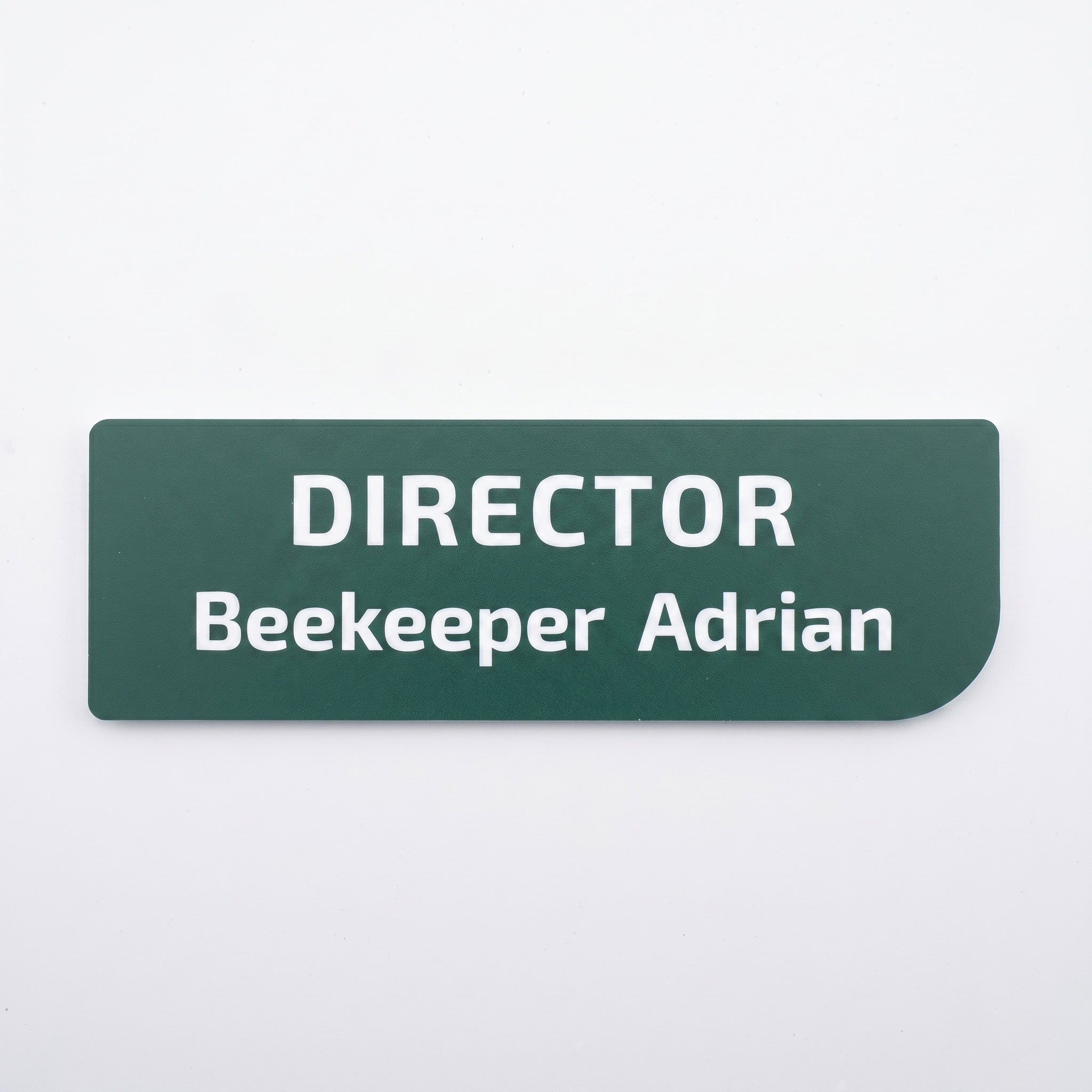 Plăcuță de identificare simplă pentru birou din acril verde închis mat, cu colțul din dreapta jos rotunjit asimetric, gravată cu textul alb „DIRECTOR Beekeeper Adrian”, pe fundal alb.