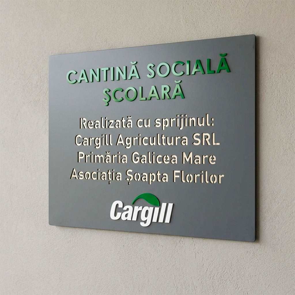 O placa de firma personalizata de culoare antracit, montata pe un perete cu textura gri, reprezentand o placa de tip mozaic pentru o „Cantina Sociala Scolara”. Imaginea pune in evidenta detaliile grafice ale produsului: textul principal si logoul Cargill sunt aplicate in relief, in timp ce lista sustinatorilor proiectului este realizata prin decupare directa in material.